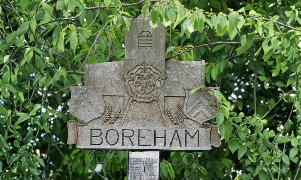 A wooden sign carved with the word "Boreham" below a rose and wheat, set against a backdrop of green leaves.