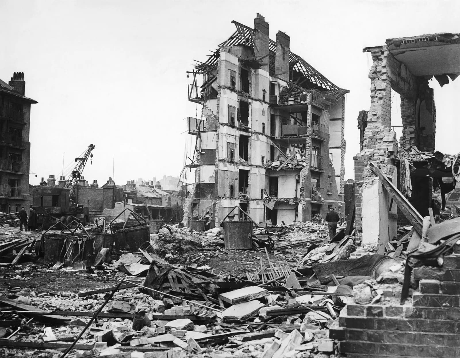 A large building with most of its walls and roof destroyed stands amidst rubble and debris, while another partially demolished structure is visible to the right and a crane operates in the background.