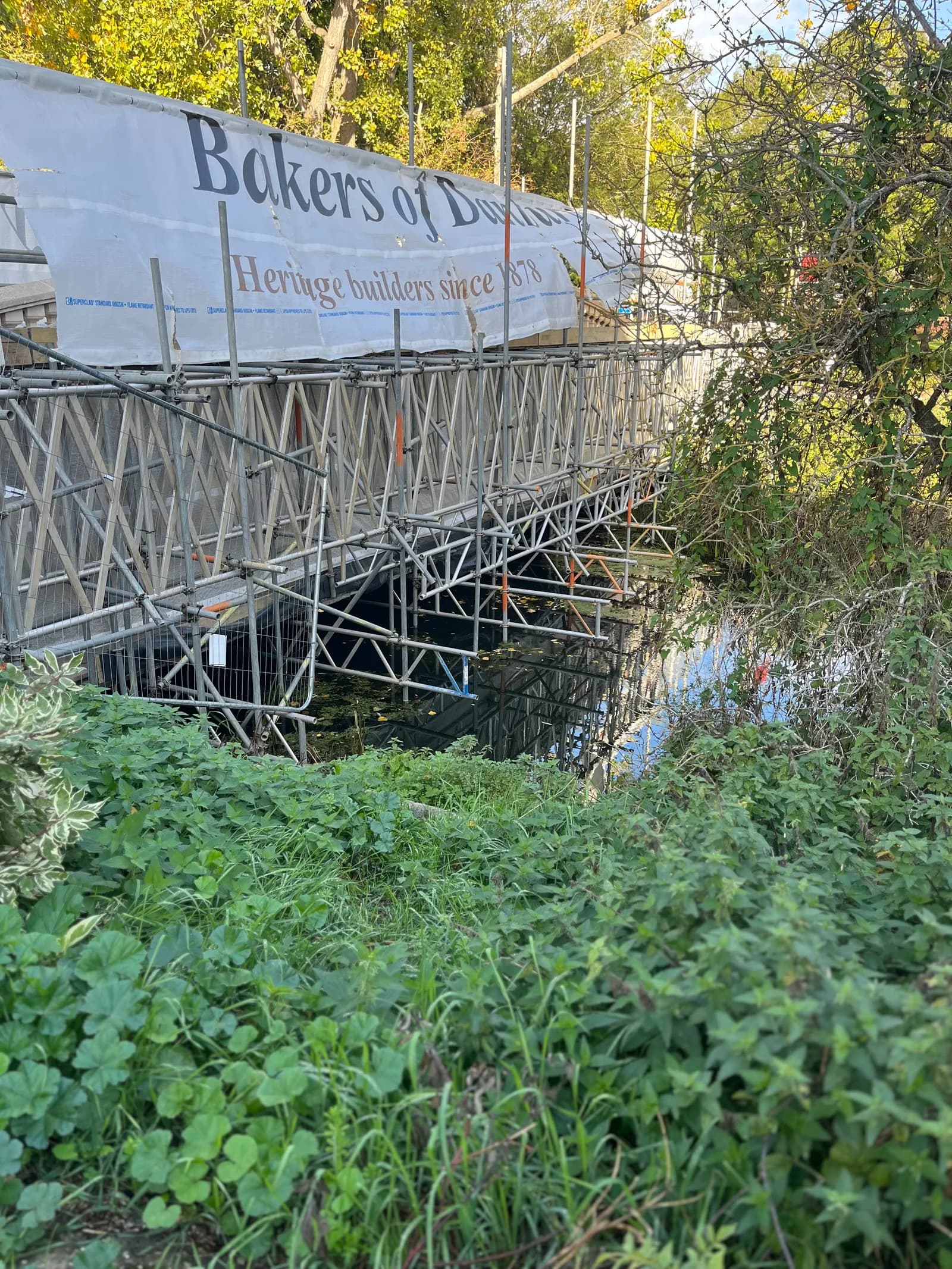 Scaffolding over water with a white banner displaying "Bakers of D... Heritage builders since 1878" surrounded by green foliage.