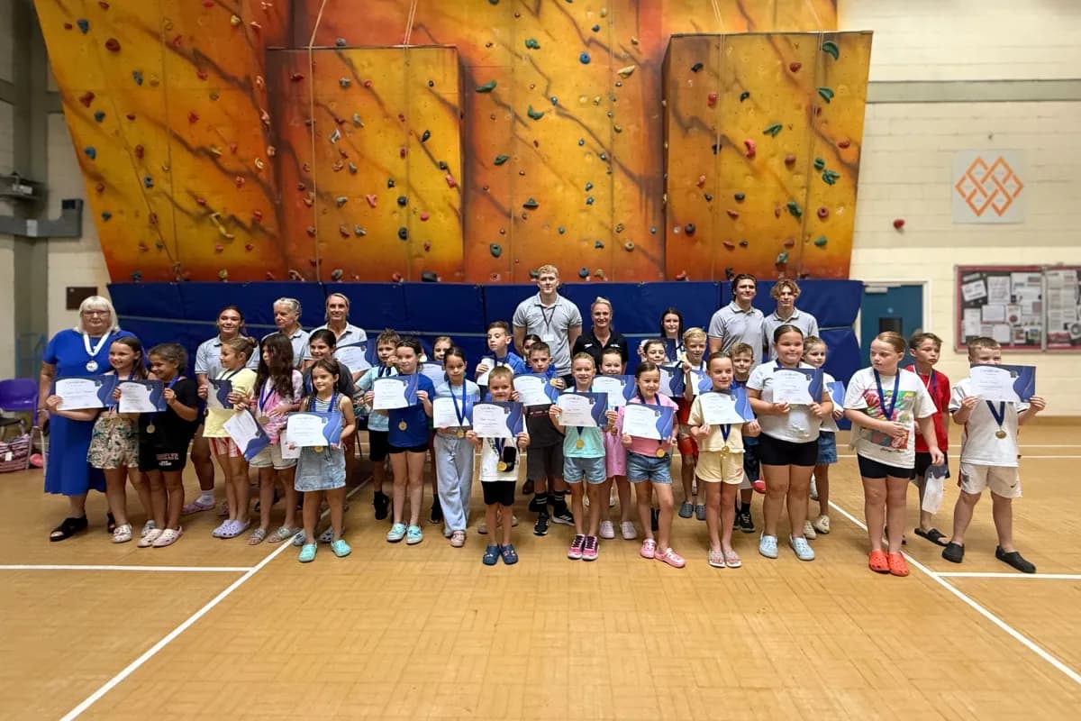 A large group of children and adults pose for a picture in front of a climbing wall, holding up certificates and wearing medals.