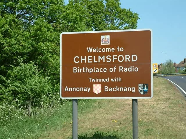A brown welcome sign announces "Welcome to CHELMSFORD Birthplace of Radio Twinning with Annonay Backnang" against a backdrop of green trees and a road.