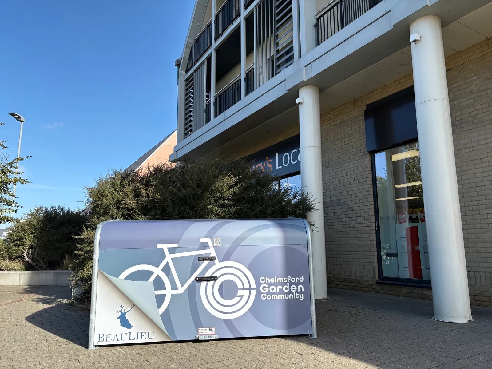 A silver bike storage unit with a blue and white design, advertising "Chelmsford Garden Community" and "Beaulieu," sits on a paved area in front of a modern building with pillars and large windows.